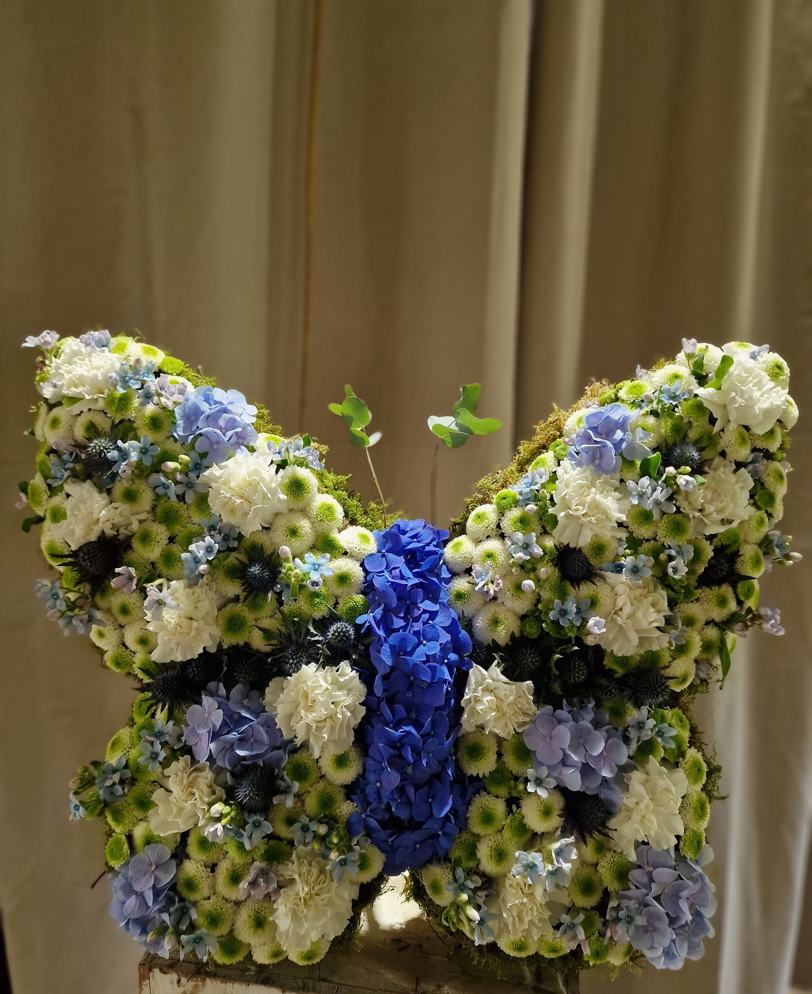 Gesteck in Schmetterlingsform aus weißen Nelken, Chrysanthemen und blauen Hortensien
