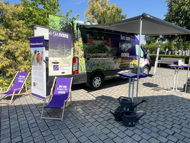 Informationsstand zur Dauergrabpflege mit Aktionsfahrzeug, Pavillon und Liegestühlen auf einem Friedhofsvorplatz