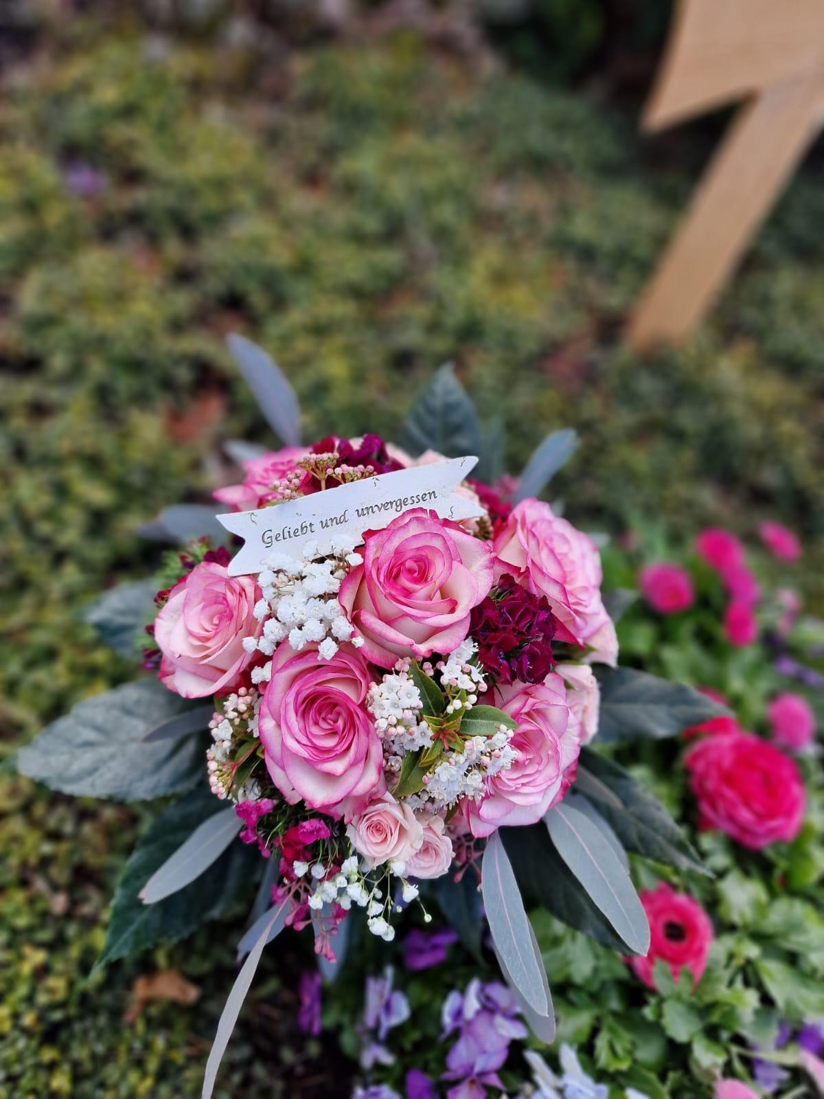 Grabgesteck aus rosafarbenen Rosen mit Schleife auf einem bepflanzten Grab